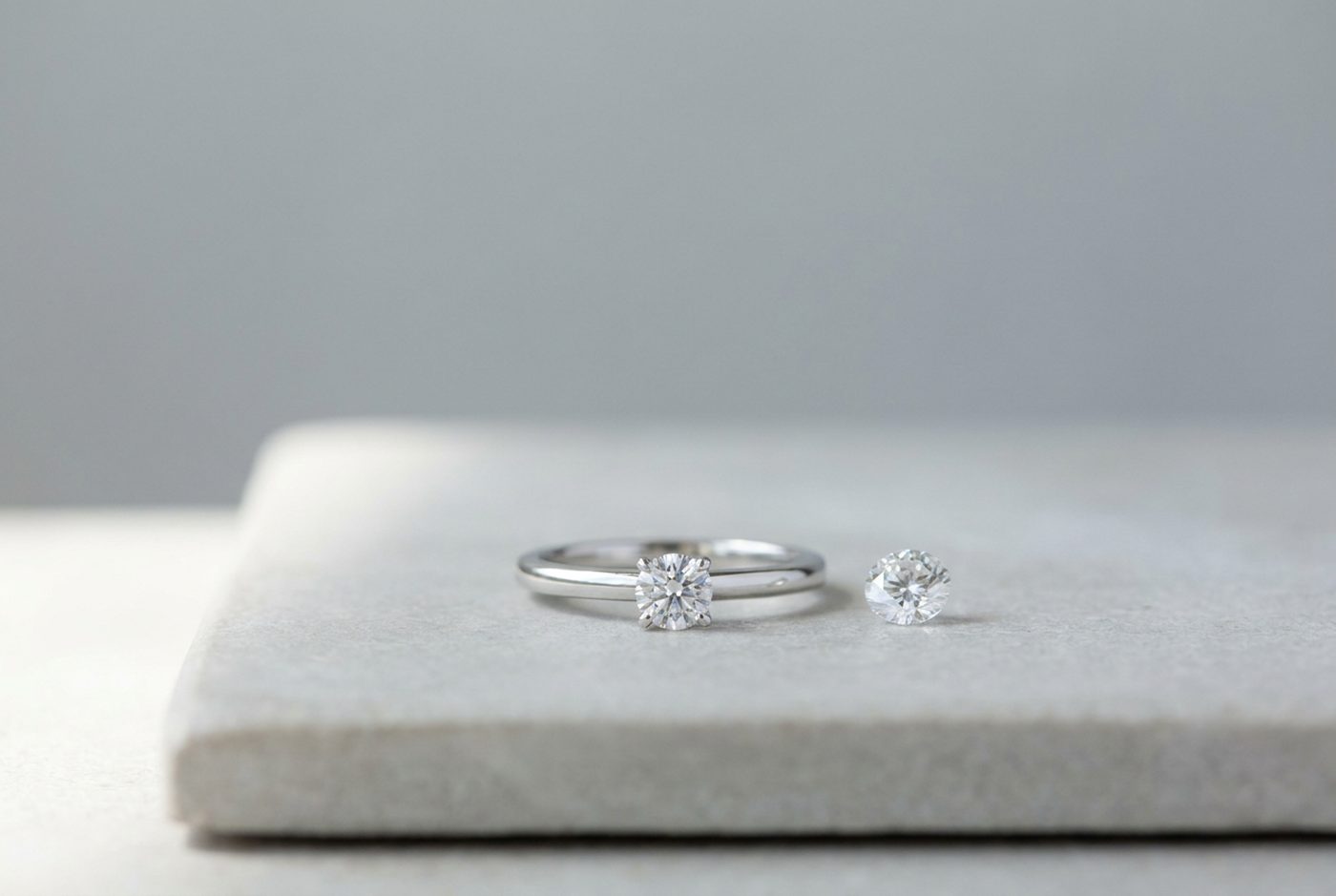 Sterling silver solitaire ring set with a round white lab stone next to a single loose round brilliant stone on a pale stone slab