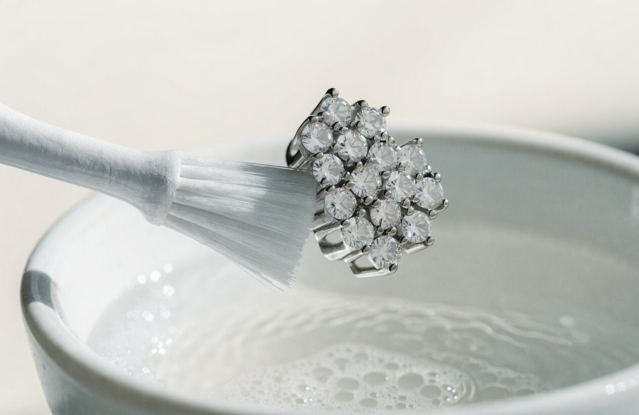 A soft toothbrush brushing the prongs of a sterling silver moissanite cluster piece over a bowl of soapy water