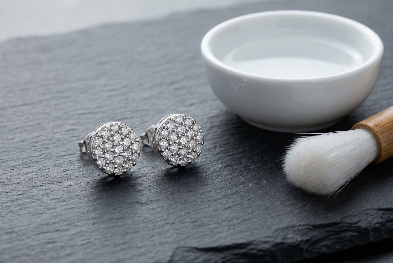 A pair of sterling silver cluster stud earrings on dark slate beside a small bowl of water and a soft brush