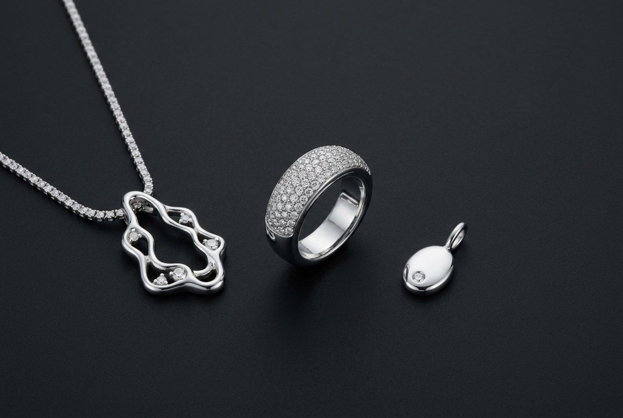 Three finished custom sterling silver moissanite pieces on a black surface: a cut-out pendant on a tennis chain, a pave ring and a small oval pendant
