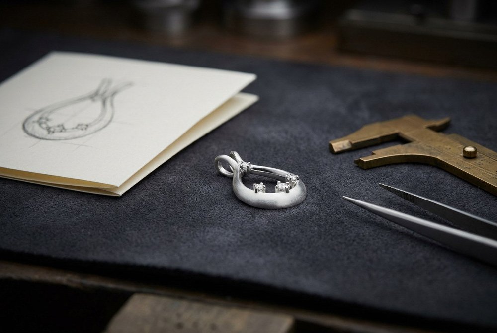 A partly set custom sterling silver pendant on a jeweler's bench beside a pencil design sketch, calipers and tweezers