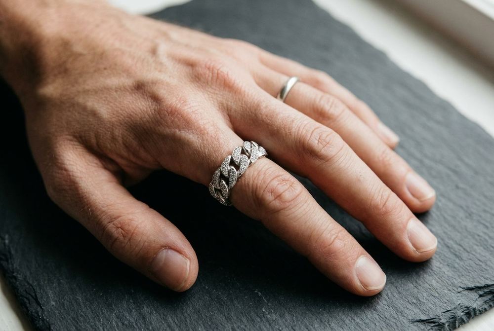 Man's hand on a slate surface wearing a stone-set sterling silver Cuban link ring and a plain silver band