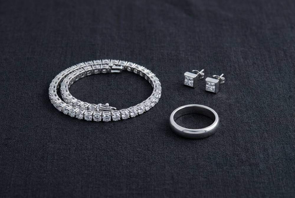 Sterling silver moissanite tennis bracelet, square stud earrings and a plain band ring arranged on dark charcoal fabric