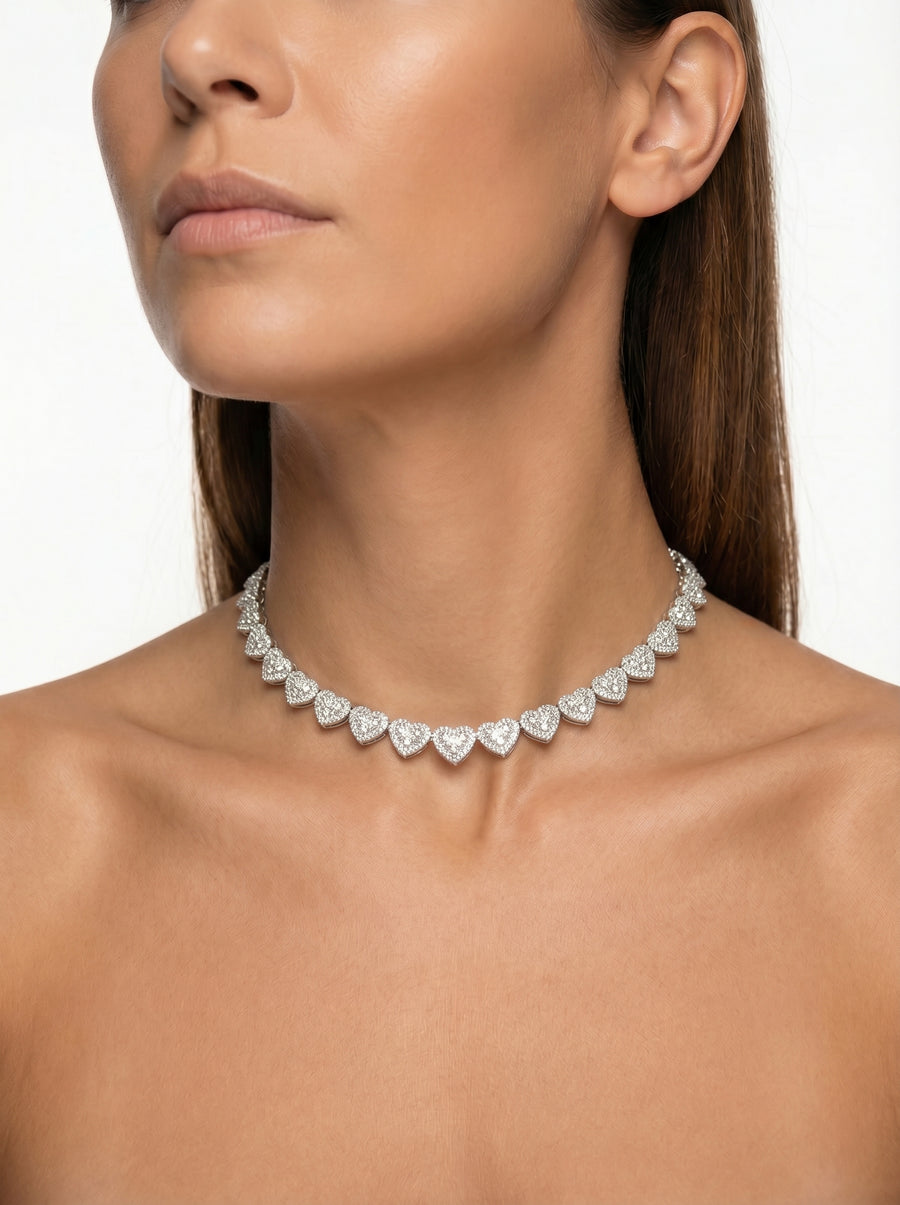 A woman with straight brown hair models the 'Pure Affection' Linked Heart Chain, a sparkling heart-shaped diamond necklace with VVS lab stones, elegantly draped on her bare neck and shoulders against a plain white background.